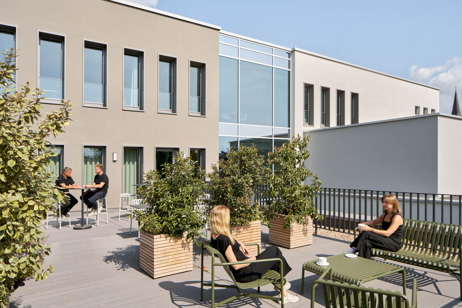 Vier Personen sitzen auf einer sonnigen Terrasse mit grünen Möbeln und Pflanzen neben einem modernen Gebäude.
