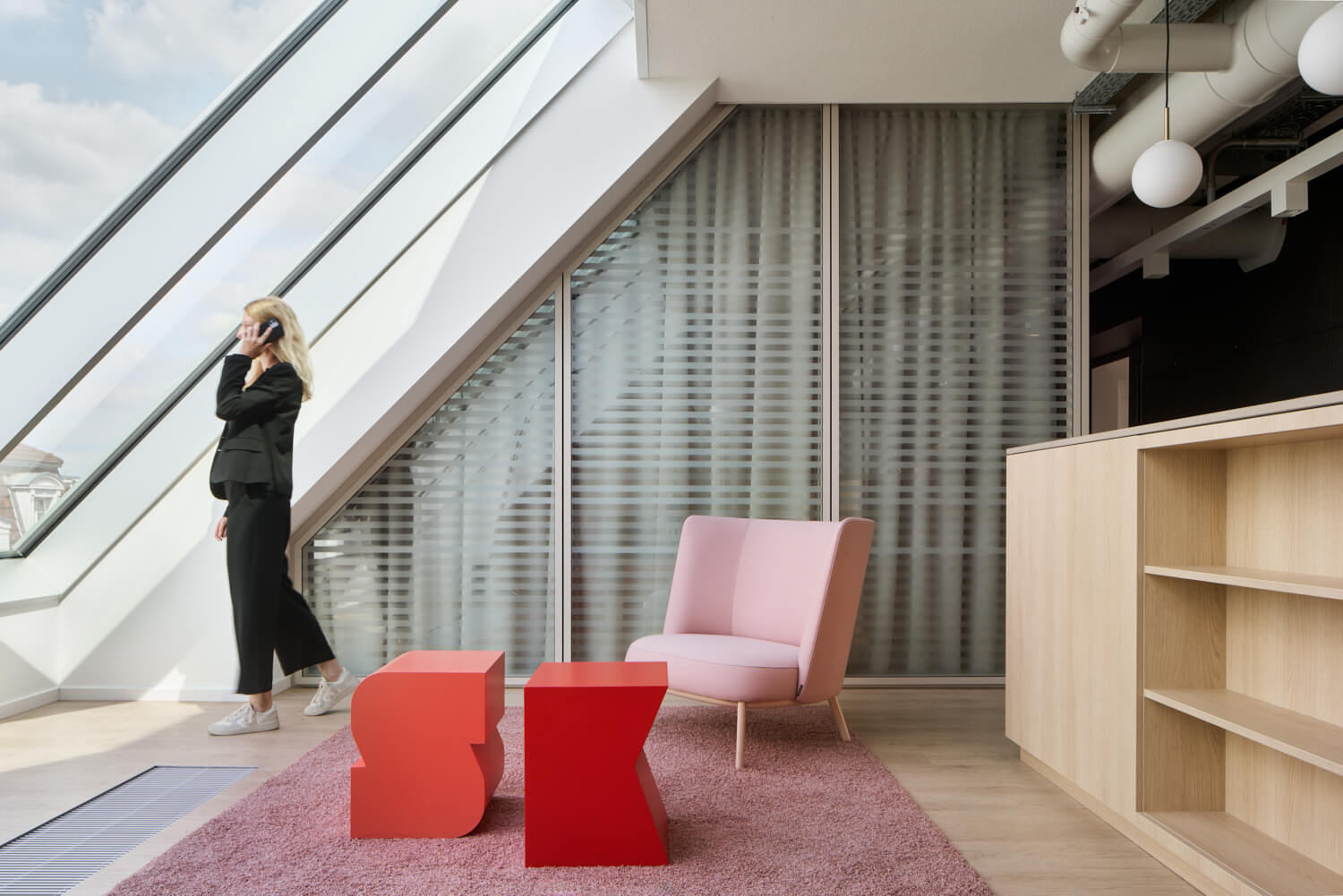 Frau im schwarzen Anzug spricht am Telefon in einem modernen Büro mit großen Dachfenstern, einem rosa Sessel und roten skulpturalen Hockern auf einem rosa Teppich.