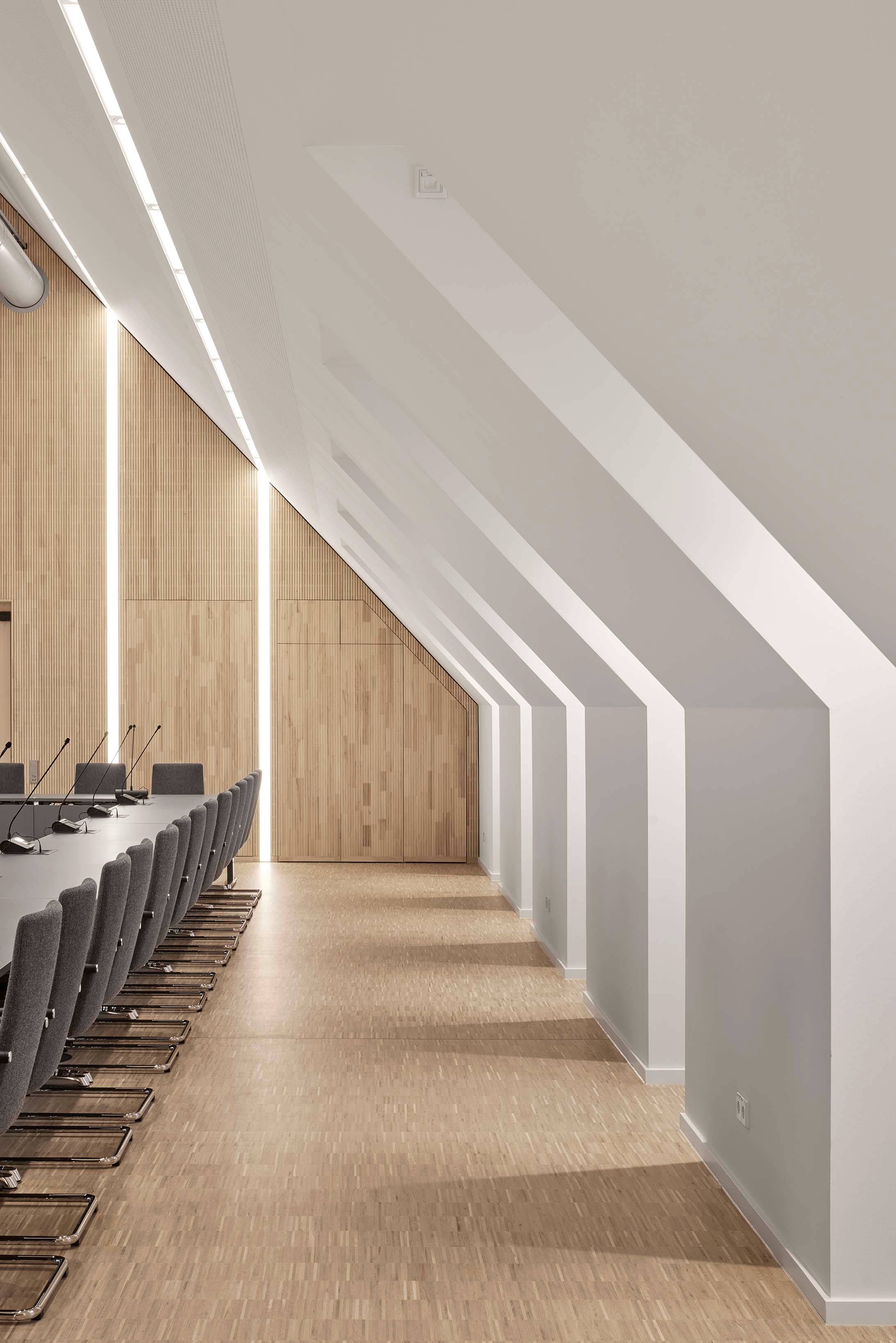 Moderner Konferenzraum mit langem Tisch, grauen Stühlen und schrägen Decken mit eingelassenen Lichtstreifen.