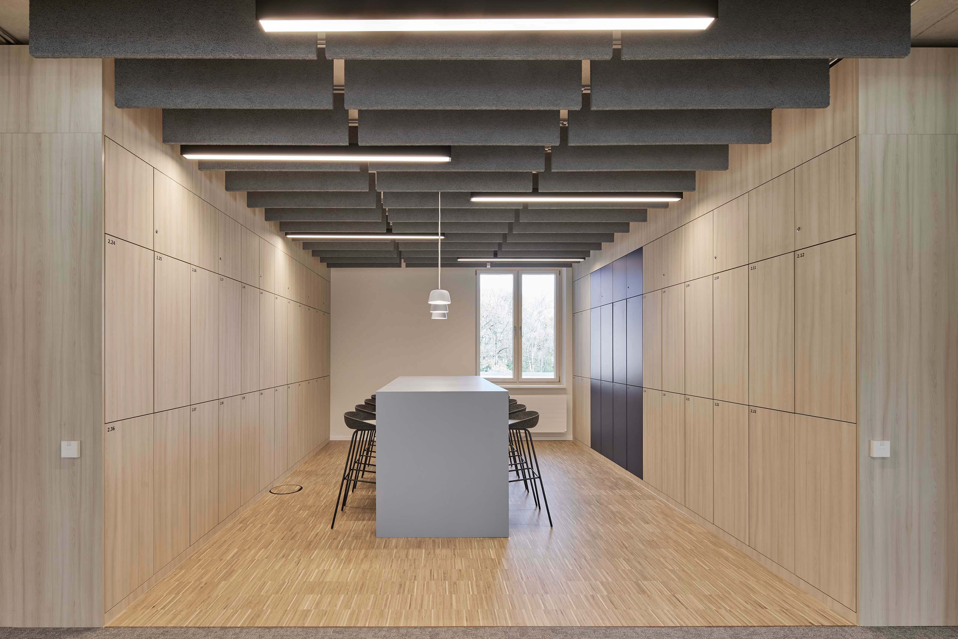 Moderner, schlichter Sitzbereich mit langem grauem Tisch, schwarzen Barhockern, mit Holz verkleideten Fächern an den Wänden und Akustikpaneelen an der Decke.