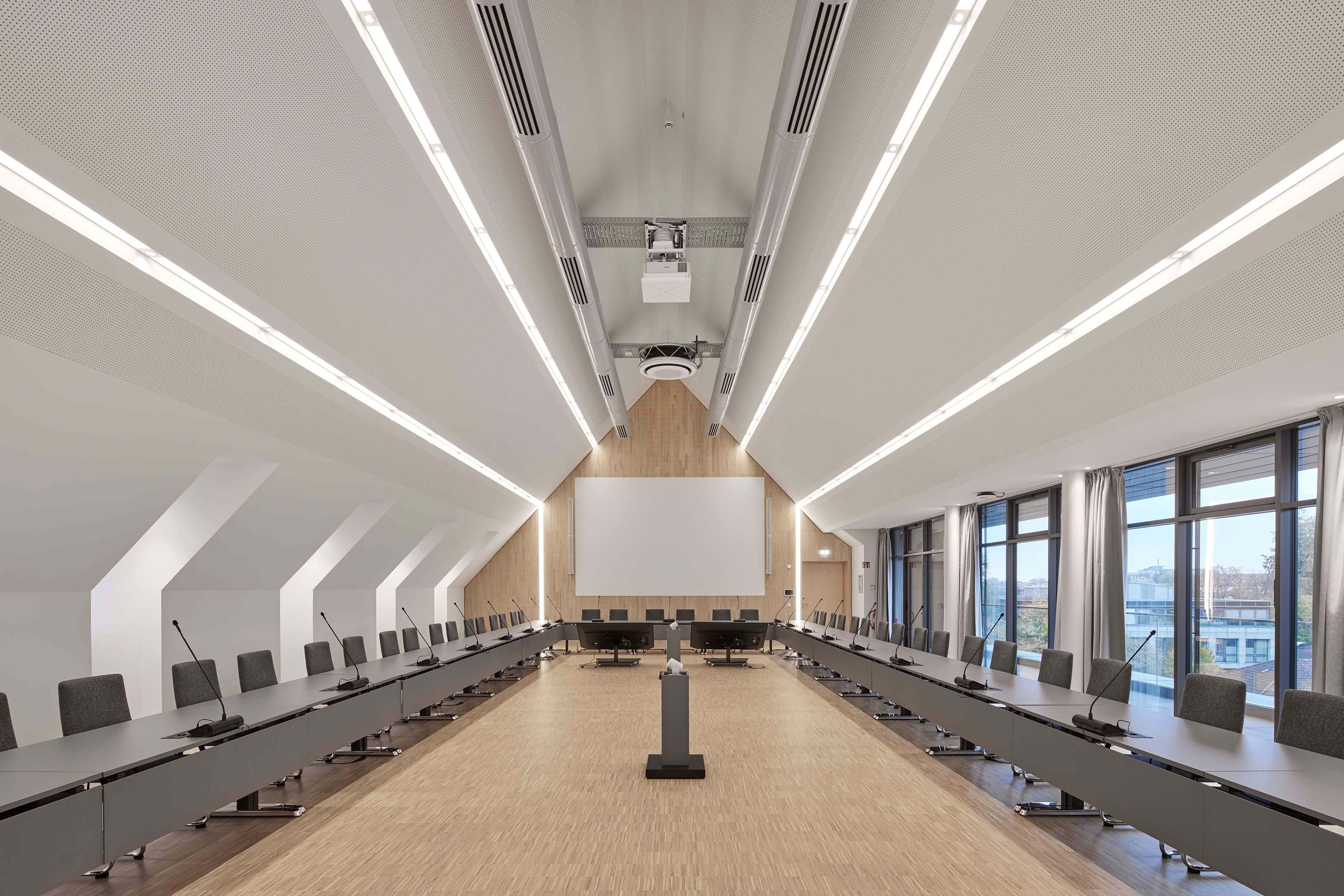 Moderner, heller Konferenzraum mit langen Tischen in U-Form, Mikrofonen auf jedem Platz und großer Leinwand an der Wand.