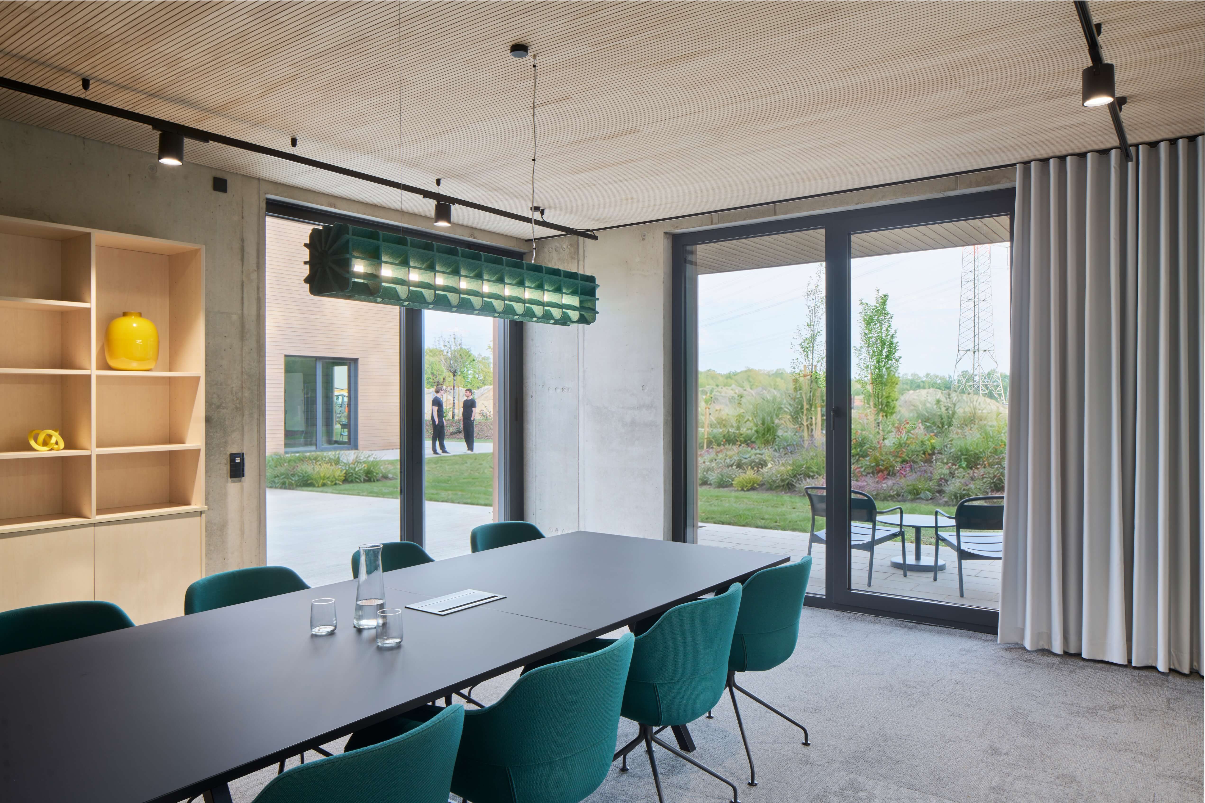 Moderner Konferenzraum mit grünem Tisch und Stühlen, großen Fenstern und Blick auf Garten.