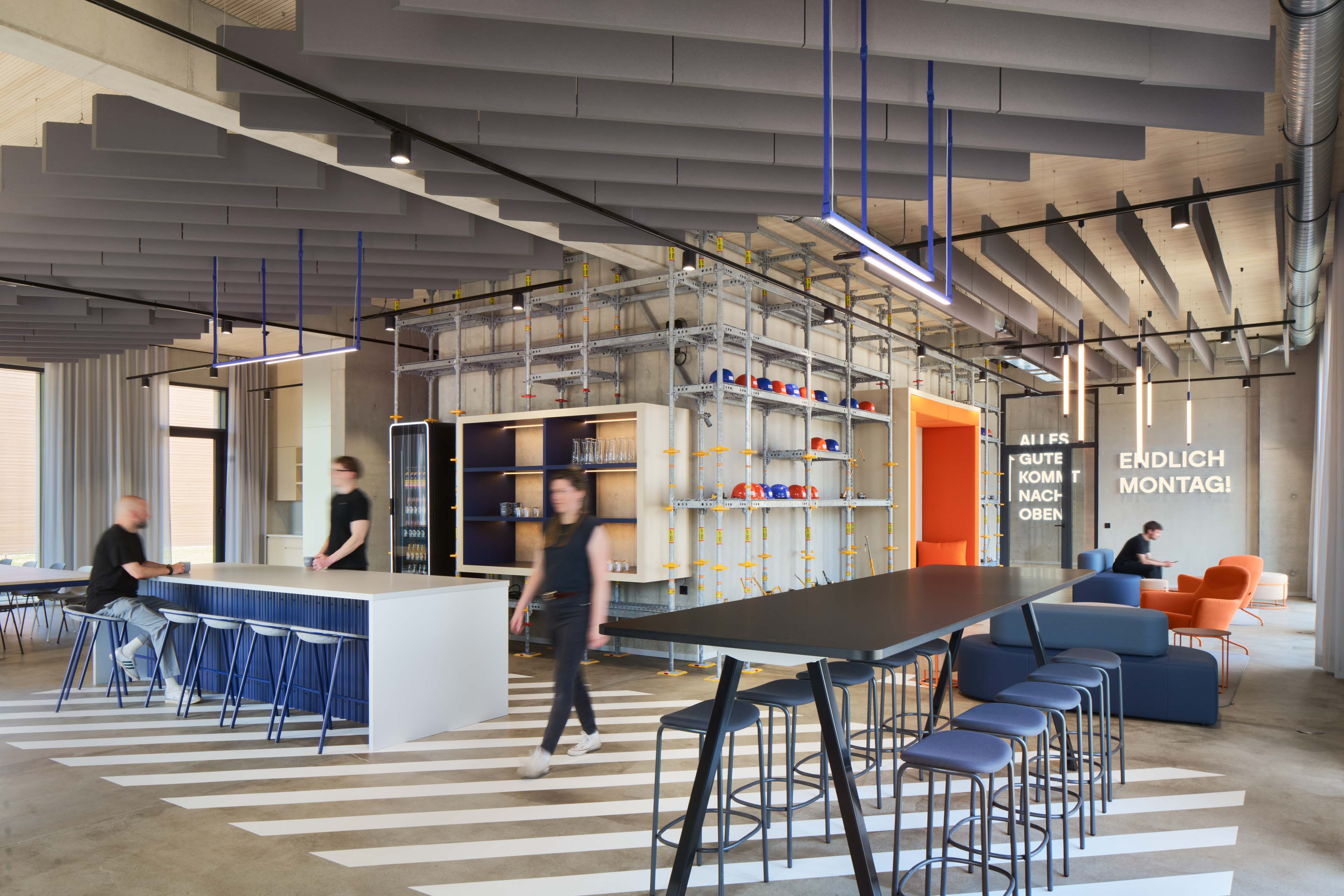 Modernes Büro mit Hochtischen, blauen Hockern, orangefarbener Sitzecke und Helmregal aus Metallgestell.