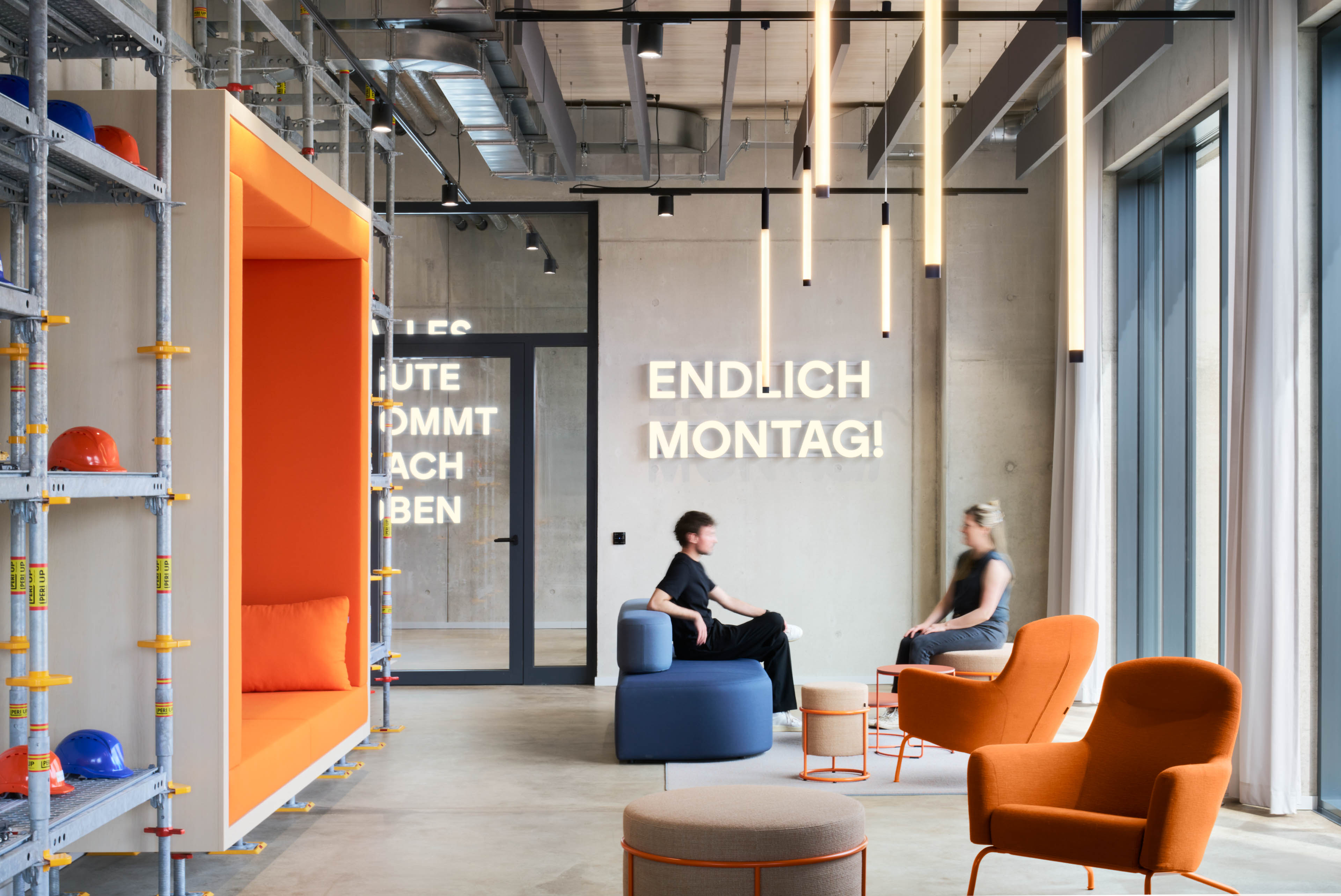 Moderne Büro-Lounge mit orangen Sesseln, blauen Sitzen, Helmregal und leuchtendem Schriftzug 'Endlich Montag!'