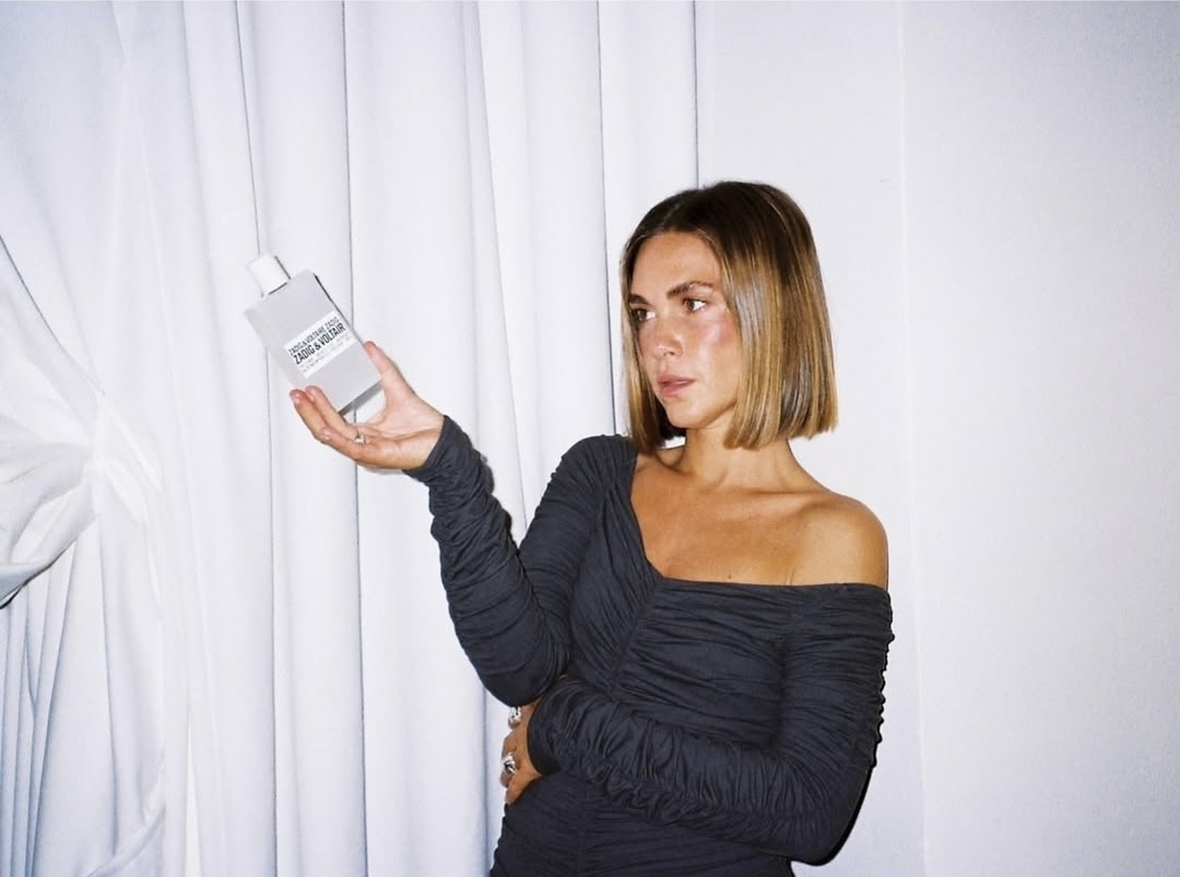 Woman with shoulder-length hair and black ruched off-shoulder dress holding a perfume bottle labeled Zadig & Voltaire.