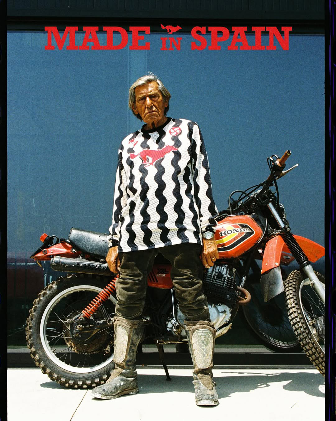 Older man wearing dark sunglasses and a navy sweater sitting at a table, holding a white gift box with a red ribbon, with text saying 'Sunnies that say "I know what I like"'.