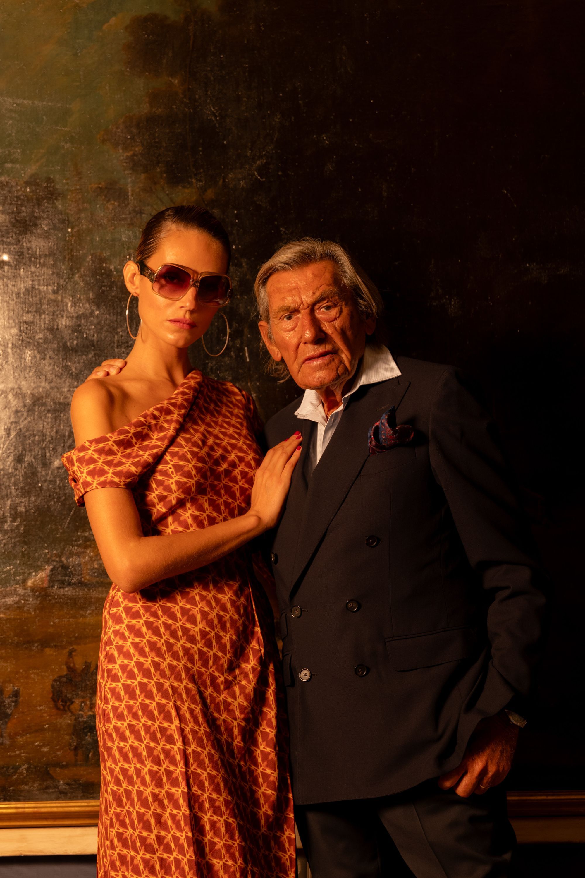 Older man wearing dark sunglasses and a navy sweater sitting at a table, holding a white gift box with a red ribbon, with text saying 'Sunnies that say "I know what I like"'.