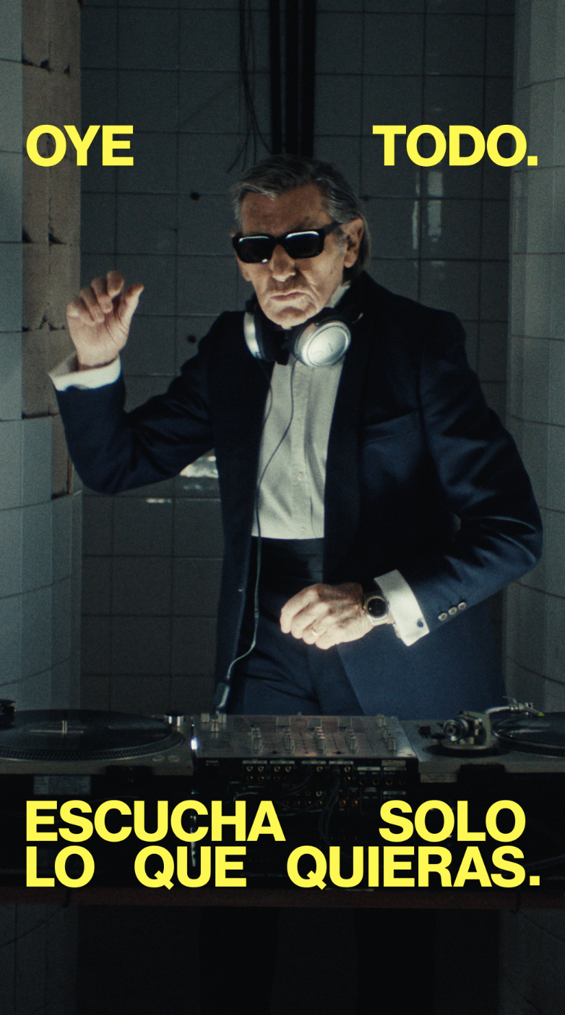 Older man wearing dark sunglasses and a navy sweater sitting at a table, holding a white gift box with a red ribbon, with text saying 'Sunnies that say "I know what I like"'.