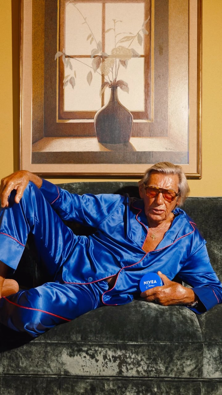 Older man wearing dark sunglasses and a navy sweater sitting at a table, holding a white gift box with a red ribbon, with text saying 'Sunnies that say "I know what I like"'.