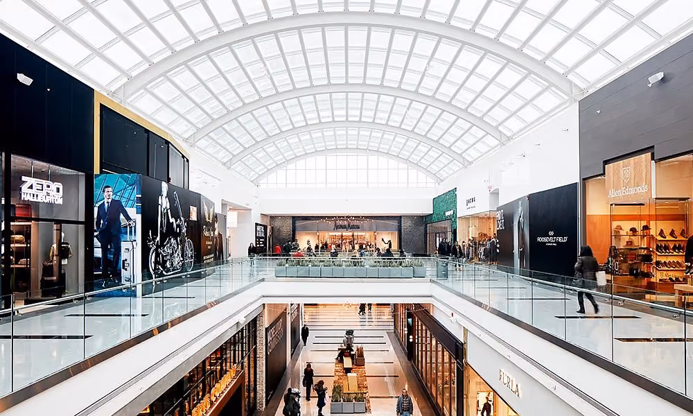Interior of a bright modern shopping mall with glass ceilings, two floors, and various retail stores.