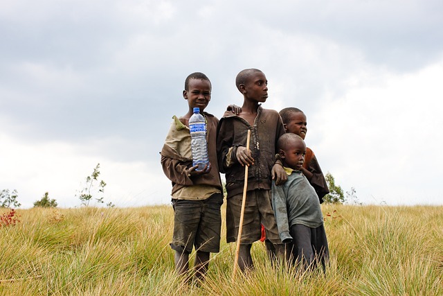 Four boys standing in a grassy field with one holding a large plastic water bottle and another holding a wooden stick.