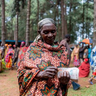 Woman in colorful patterned clothing holding money outdoors with people and trees in the background.