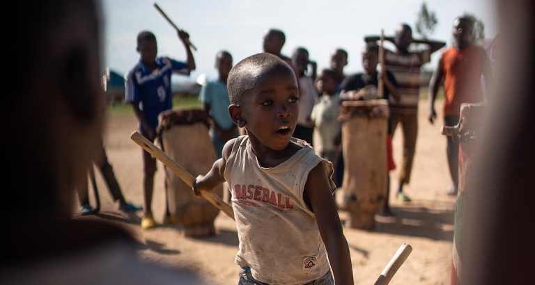 Young boy in a sleeveless shirt holding sticks, playing drums outdoors with a group of children in the background.