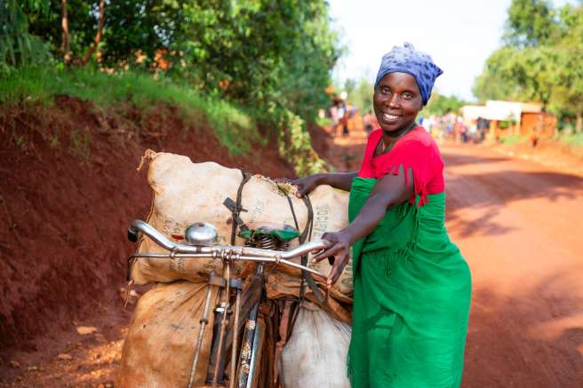 Smiling woman wearing a red shirt and green dress stands beside a bicycle loaded with large sacks on a dirt road.