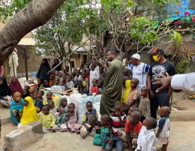 A group of children and adults gathered outdoors around bags of supplies in a community setting with trees and buildings in the background.
