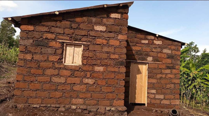 Small rustic house made of red and brown bricks with a wooden door and window, surrounded by vegetation under a clear sky.