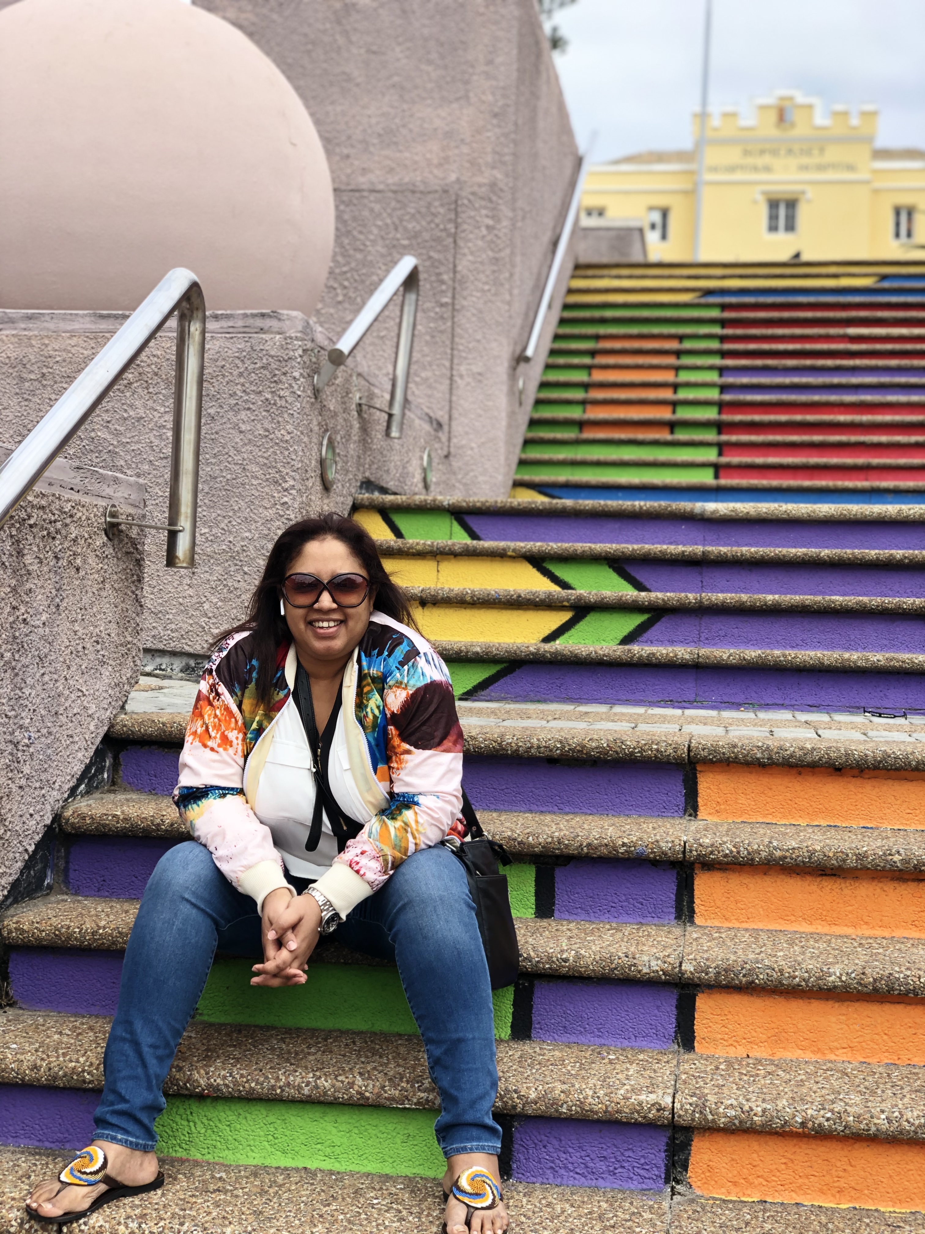 Woman wearing sunglasses and colorful jacket sitting on vibrant green, purple, orange, and yellow painted stairs with metal railings.