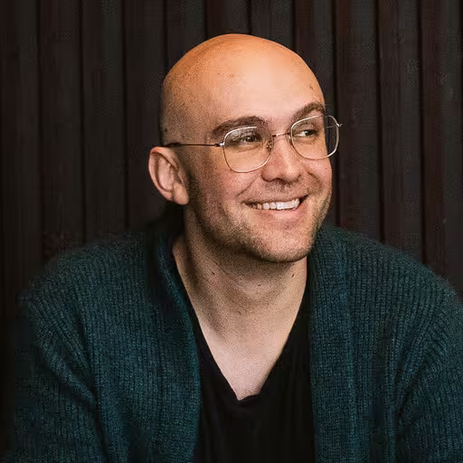 Jared smiling with glasses wearing a dark green sweater and black shirt against a dark background.