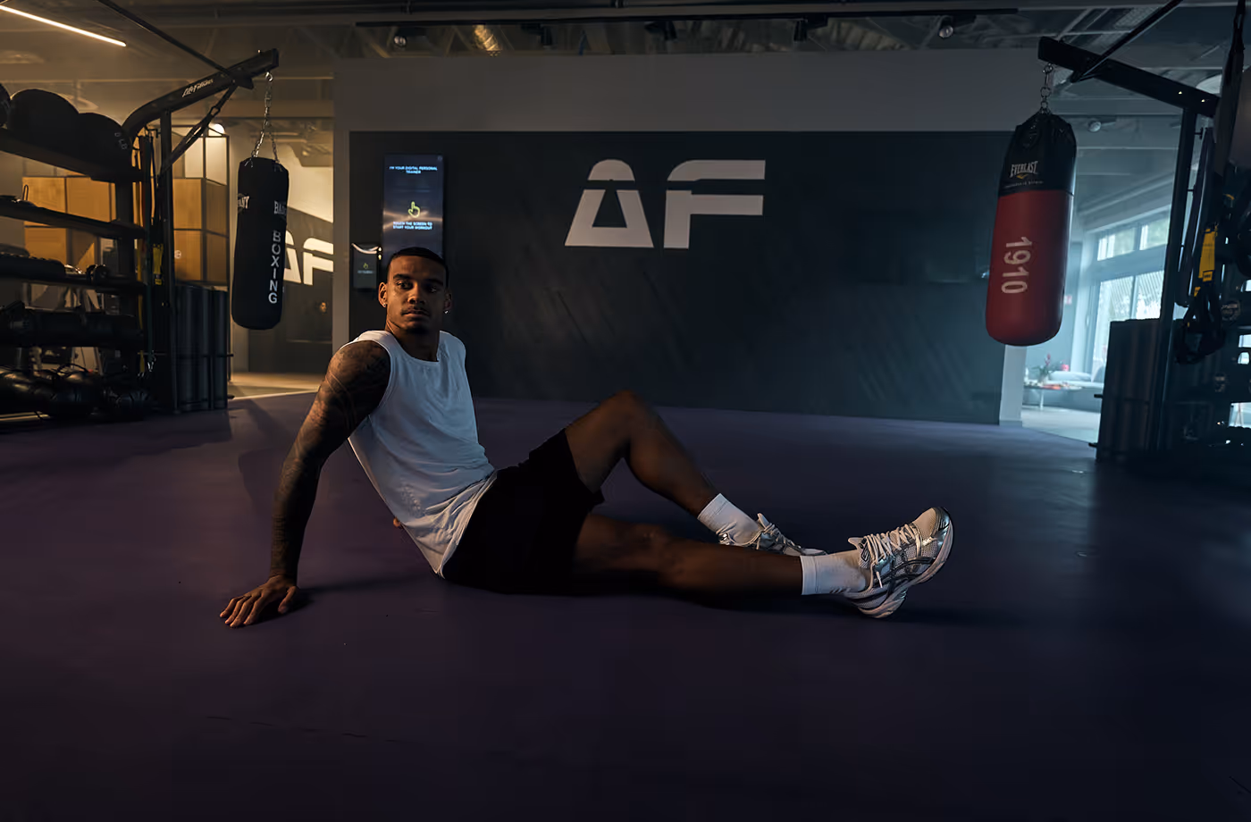 Man in white tank top and black shorts sitting on the floor of a boxing gym with punching bags and gym equipment.