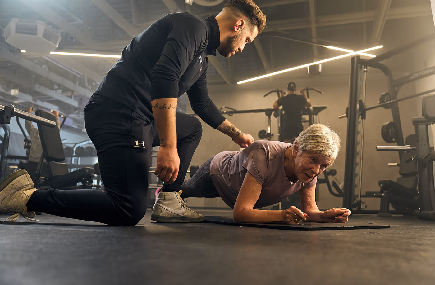 Trainer assists an elderly woman doing a plank exercise on a mat in a gym.