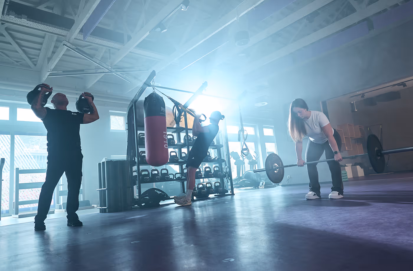 Three people exercising in a gym with kettlebells, suspension straps, and a barbell.