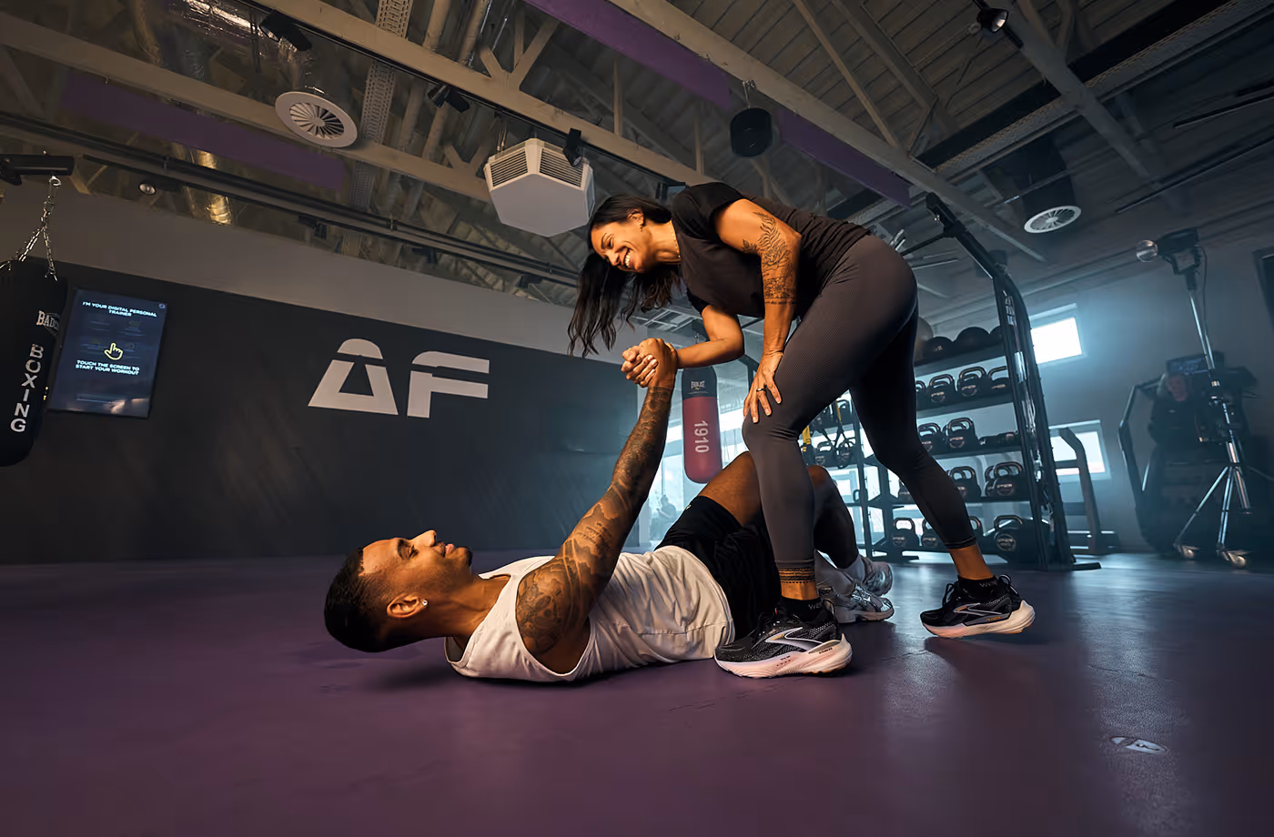 Woman helping man up with a handshake on a purple gym floor inside a fitness center.
