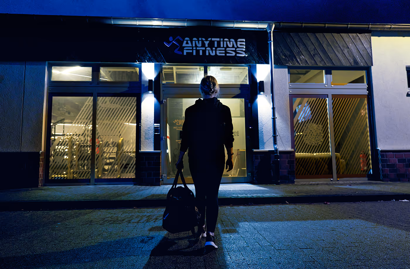 Person carrying a gym bag walking towards the entrance of an Anytime Fitness gym at night.