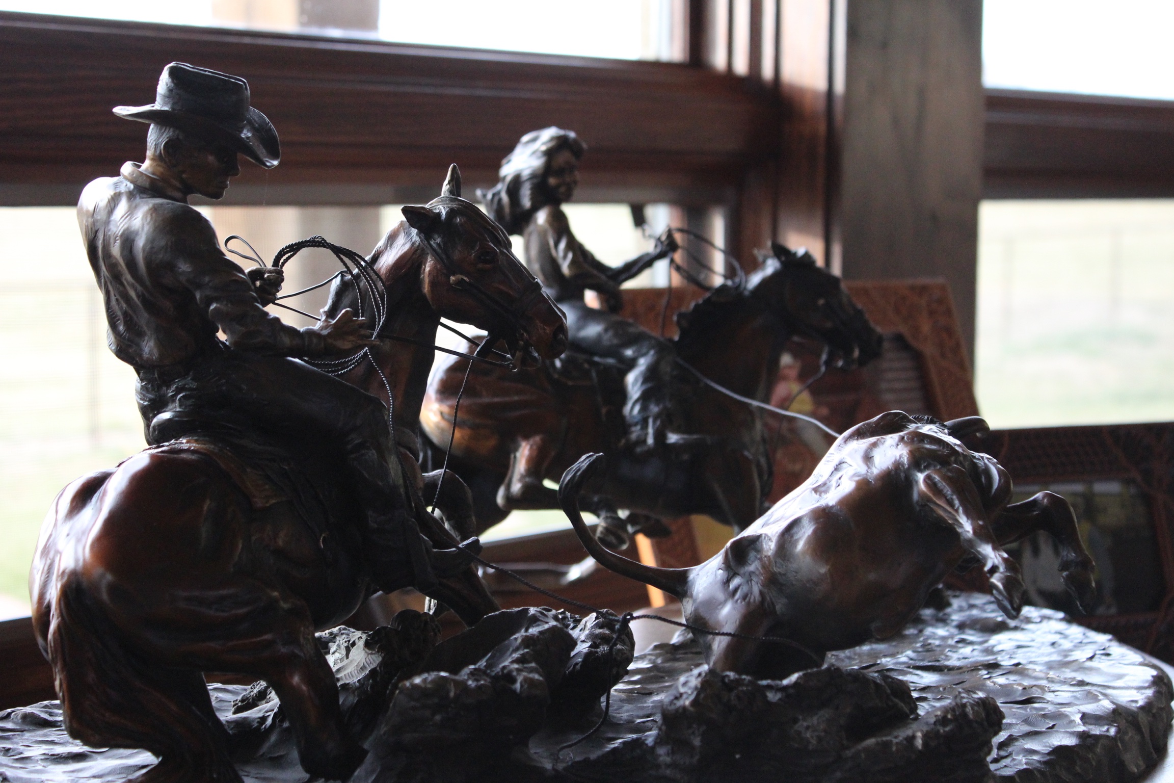 A bronze sculpture depicts two cowboys on horseback roping a steer, captured mid-action with lassos extended, displayed indoors near a large window.