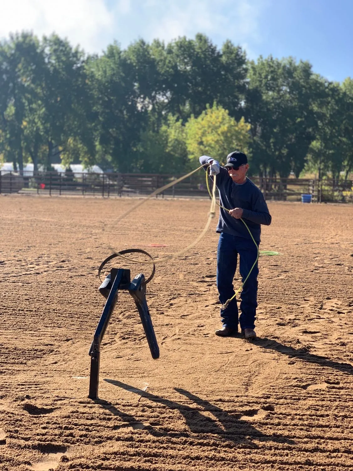 An instructor in a cap and sunglasses swings a rope at a ground roping dummy in a sandy arena