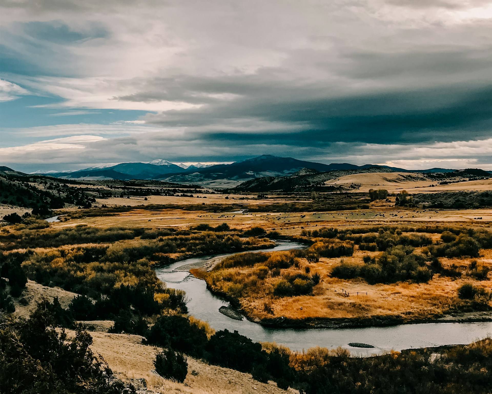 A river winds through golden autumn grassland and willows toward snow-dusted mountains under a dramatic, cloudy sky
