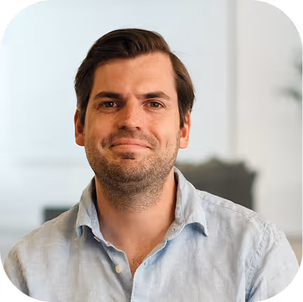 Smiling man with dark hair wearing a light blue button-up shirt in a bright, blurred indoor setting.