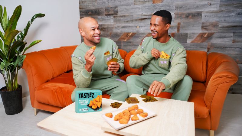 Two people wearing the custom green TAST!EZ sweatsuits play cards on an orange couch