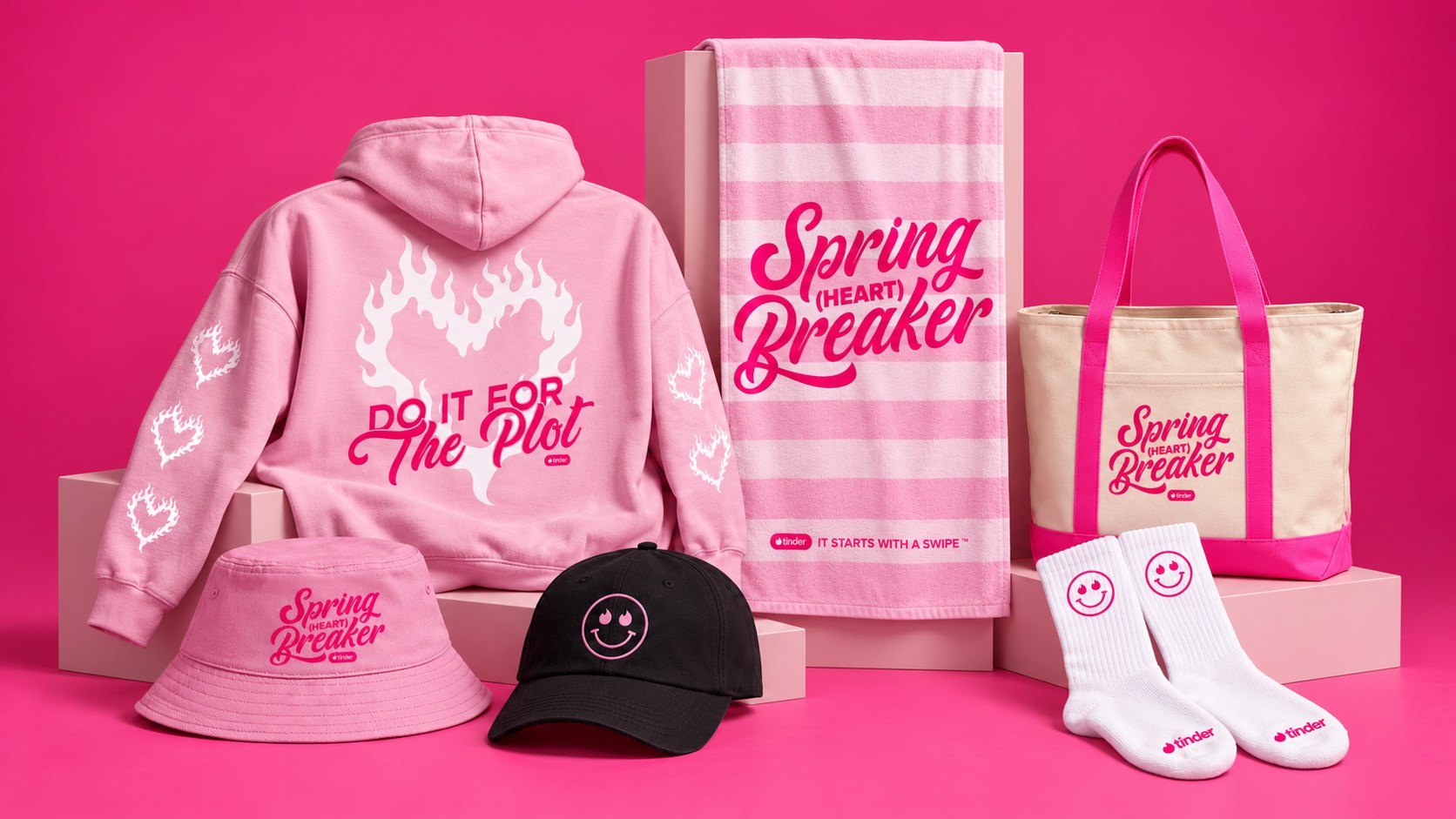 Tinder custom merchandise on a pink studio set: pink hoodie, striped beach towel, Spring Heart Breaker tote and bucket hat, smiley cap and socks