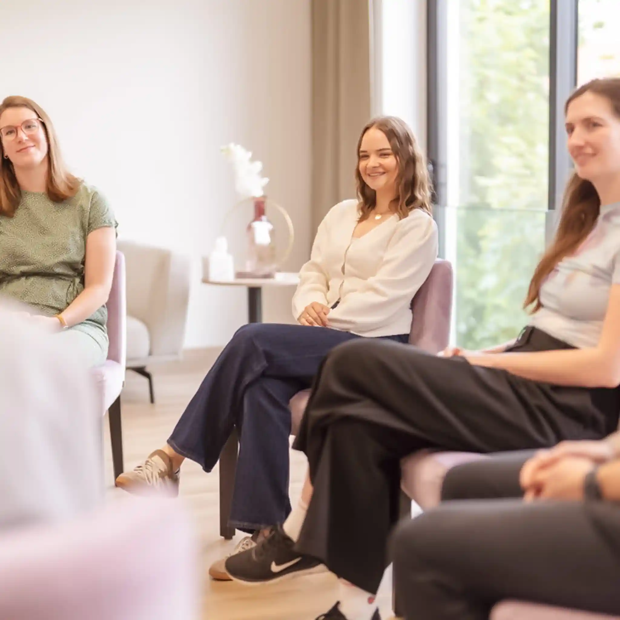 Mehrere Patientinnen sitzen in einem Kreis auf gepolsterten Stühlen und blicken gespannt auf eine weitere Person im Sitzkreis.