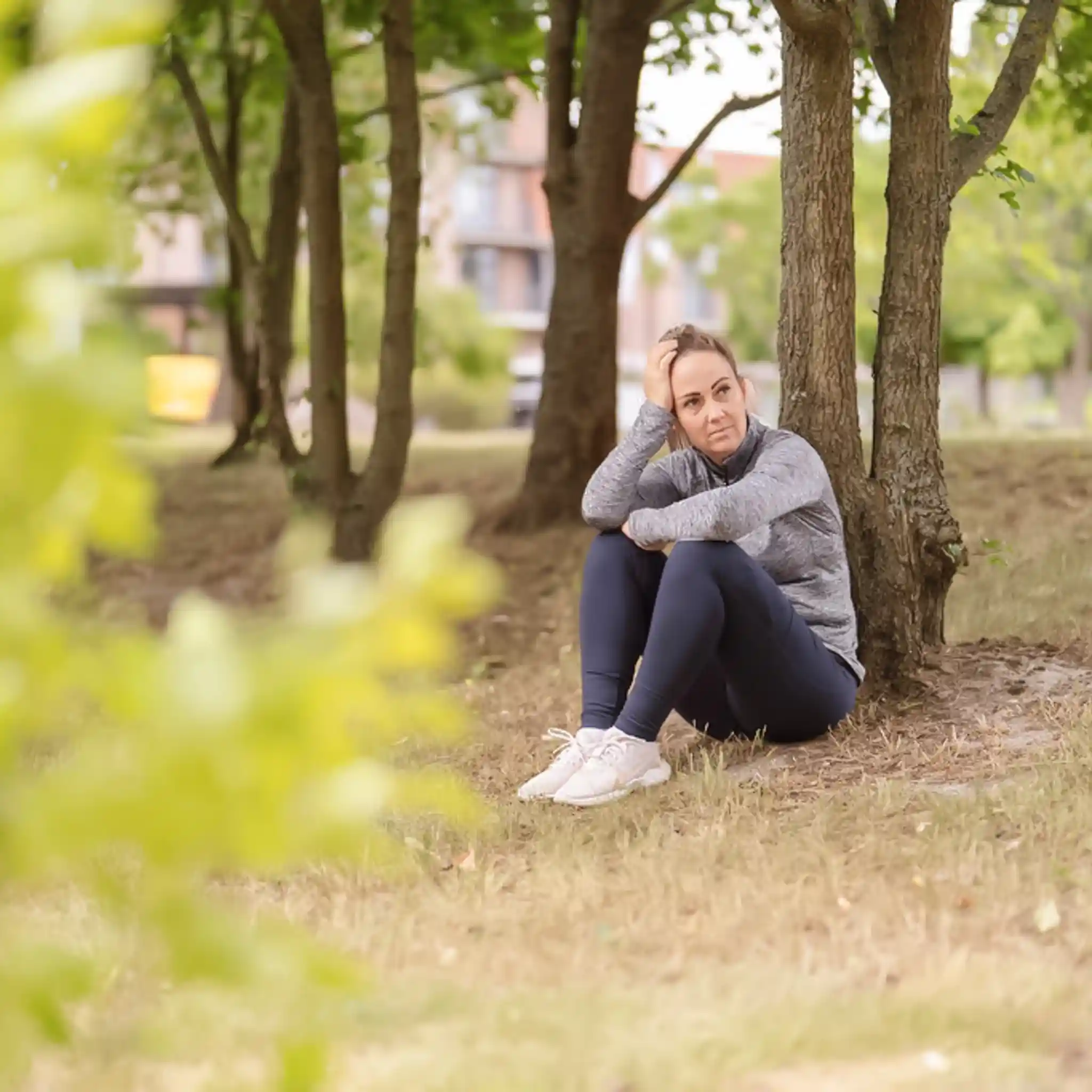 Eine Frau in Trainingsklamotten sitzt an einen Baum gelehnt auf dem Boden und blickt in die Ferne.