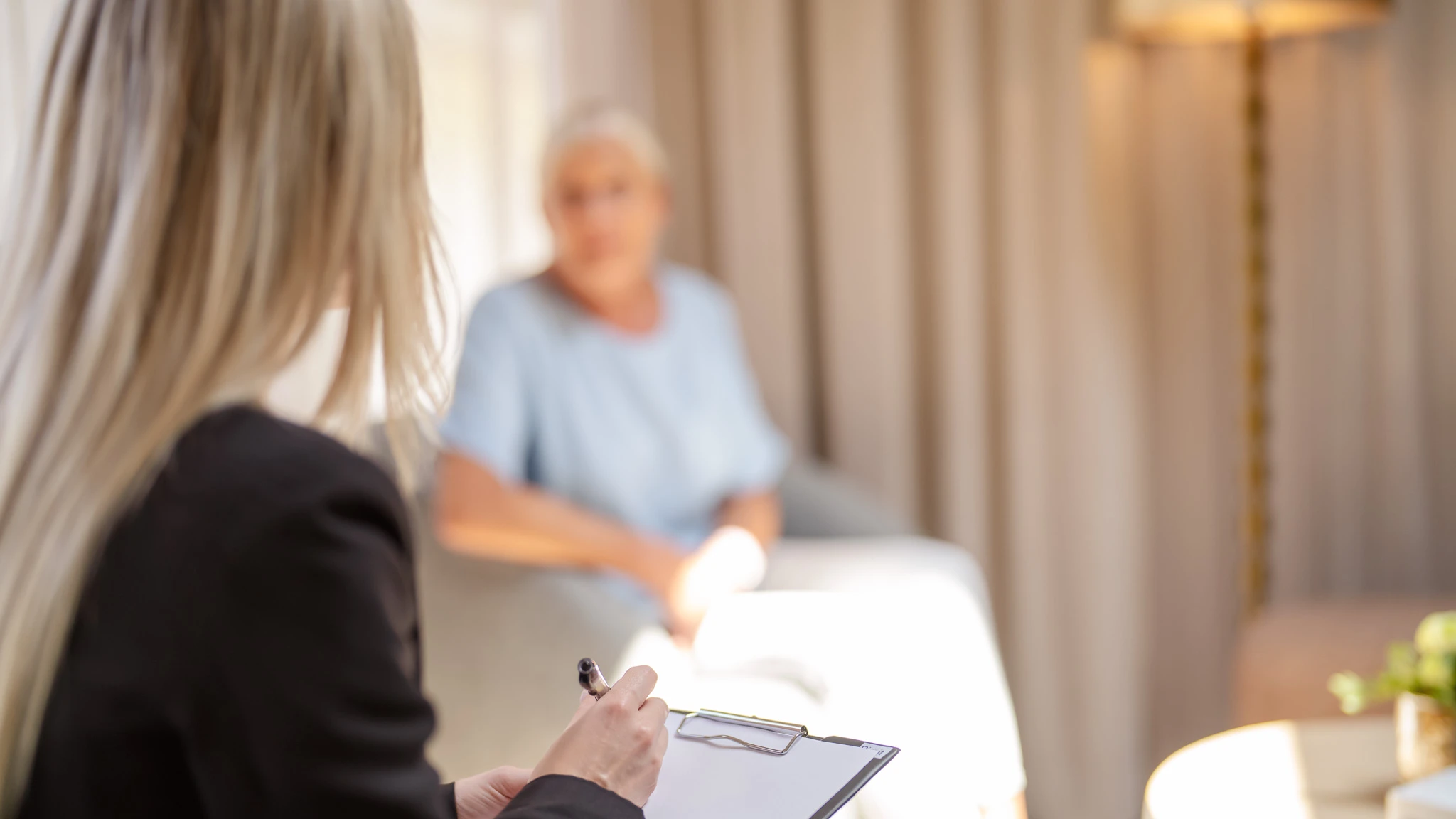 Eine Therapeutin mit Klemmbrett im Gespräch mit einer Patientin. Sie ist im Fokus und sitzt mit dem Rücken zur Kamera.
