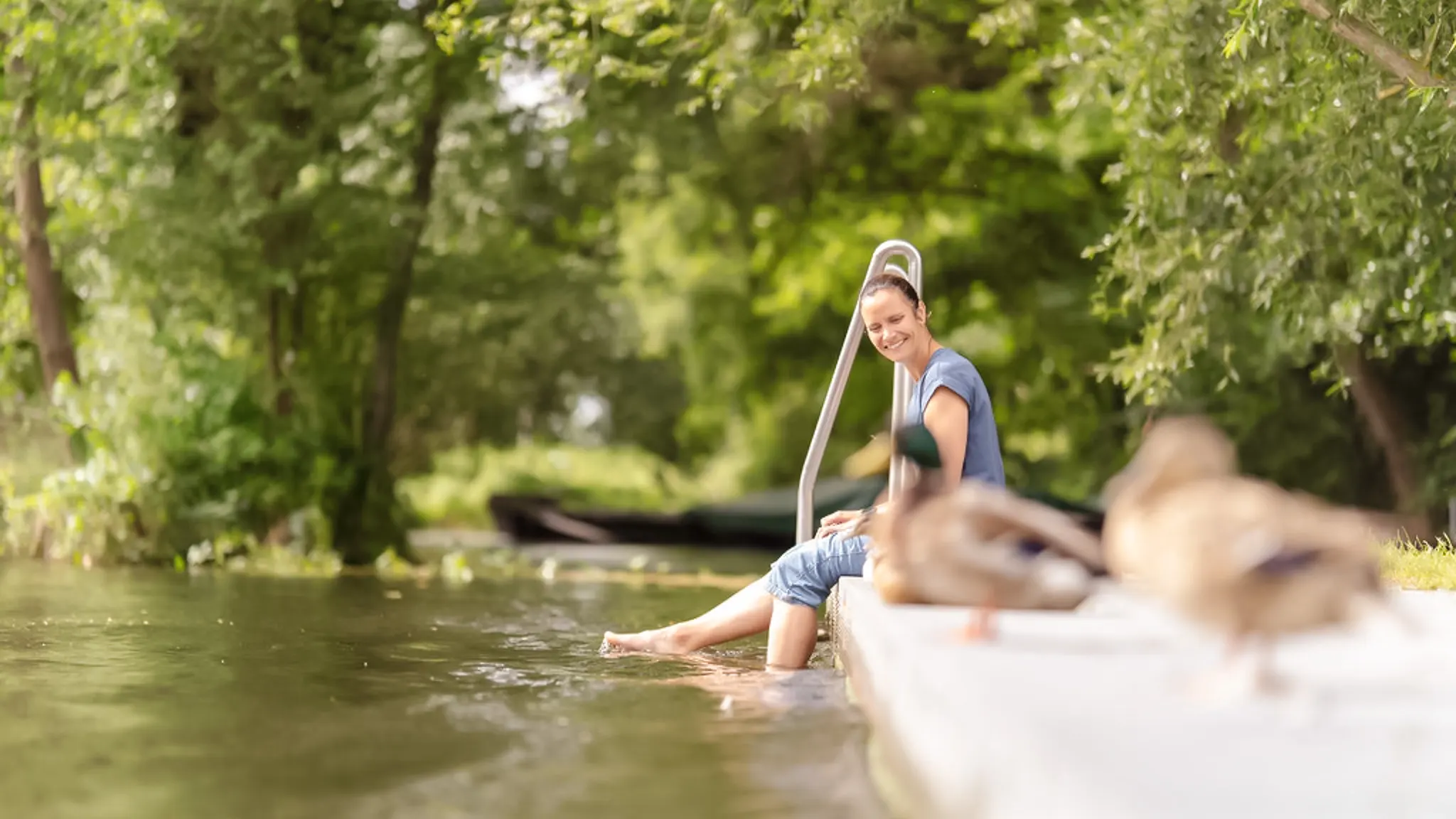 Eine Frau sitzt am Rand eines Beckens und lässt die Beine ins Wasser hängen. Lachend beobachtet sie zwei Enten.
