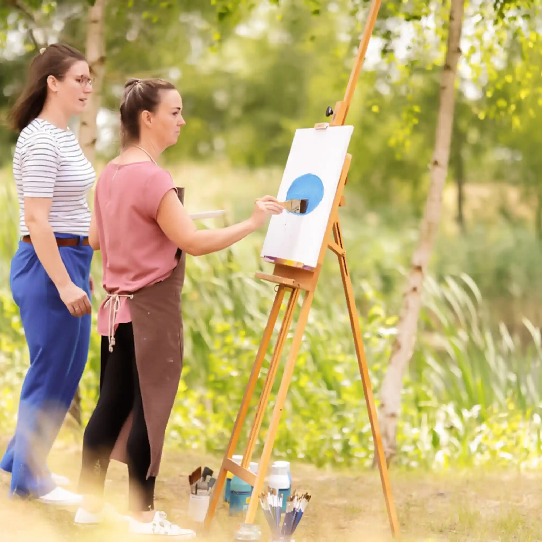 Eine Patientin malt ein Bild auf einer Leinwand. Sie ist draußen im Grünen. Neben ihr steht eine Kunsttherapeutin.
