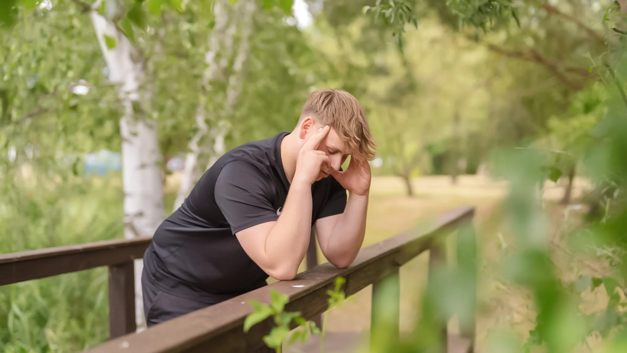 Ein Mann stützt sich mit den Ellenbogen auf dem Geländer einer kleinen Holzbrücke im Wald ab und hat beide Hände an seine Schläfen gelegt.