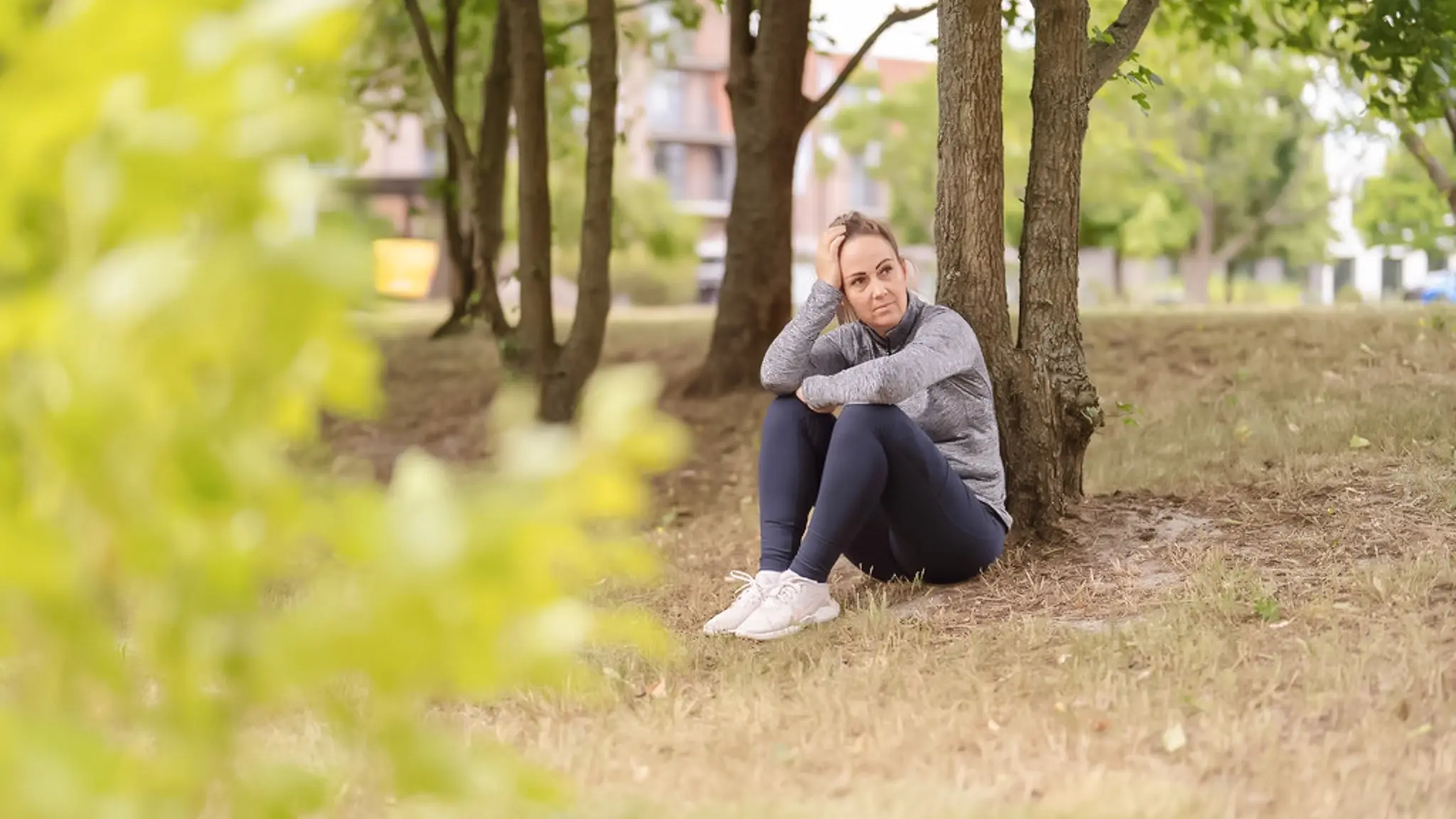 Eine Frau in Trainingsklamotten sitzt auf dem Boden, an einen Baum gelehnt. Sie blickt zur Seite und hat ihren Kopf auf ihre Hand gestützt.