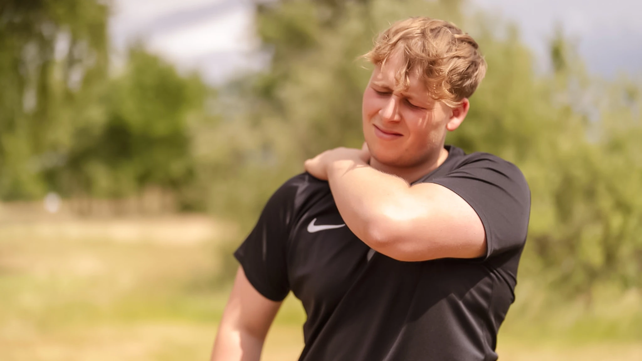 Ein junger Mann in einem Sportshirt steht in der Natur und hält sich mit schmerzerfülltem Blick die rechte Schulter.