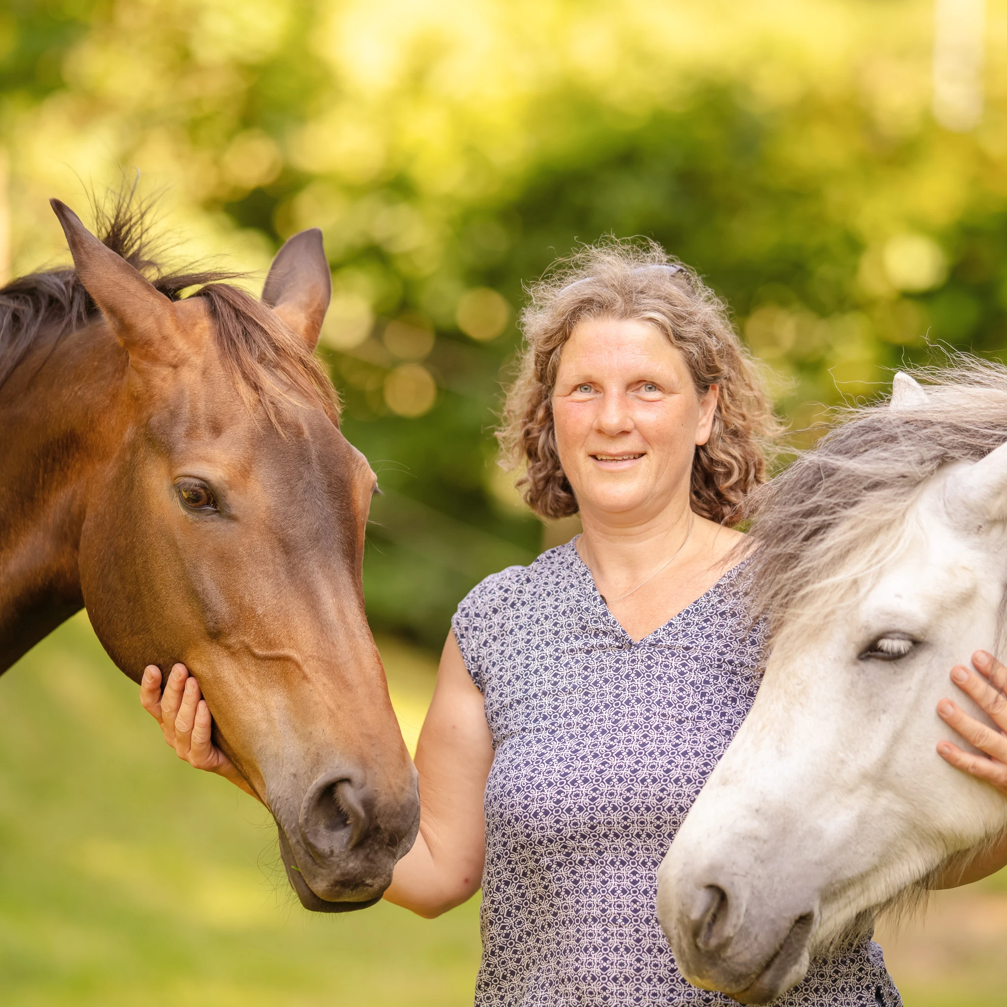 Eine Patientin streichelt die Köpfe eines Pferdes und eines Ponys bei der Pferdetherapie. Dabei lächelt sie in die Kamera.