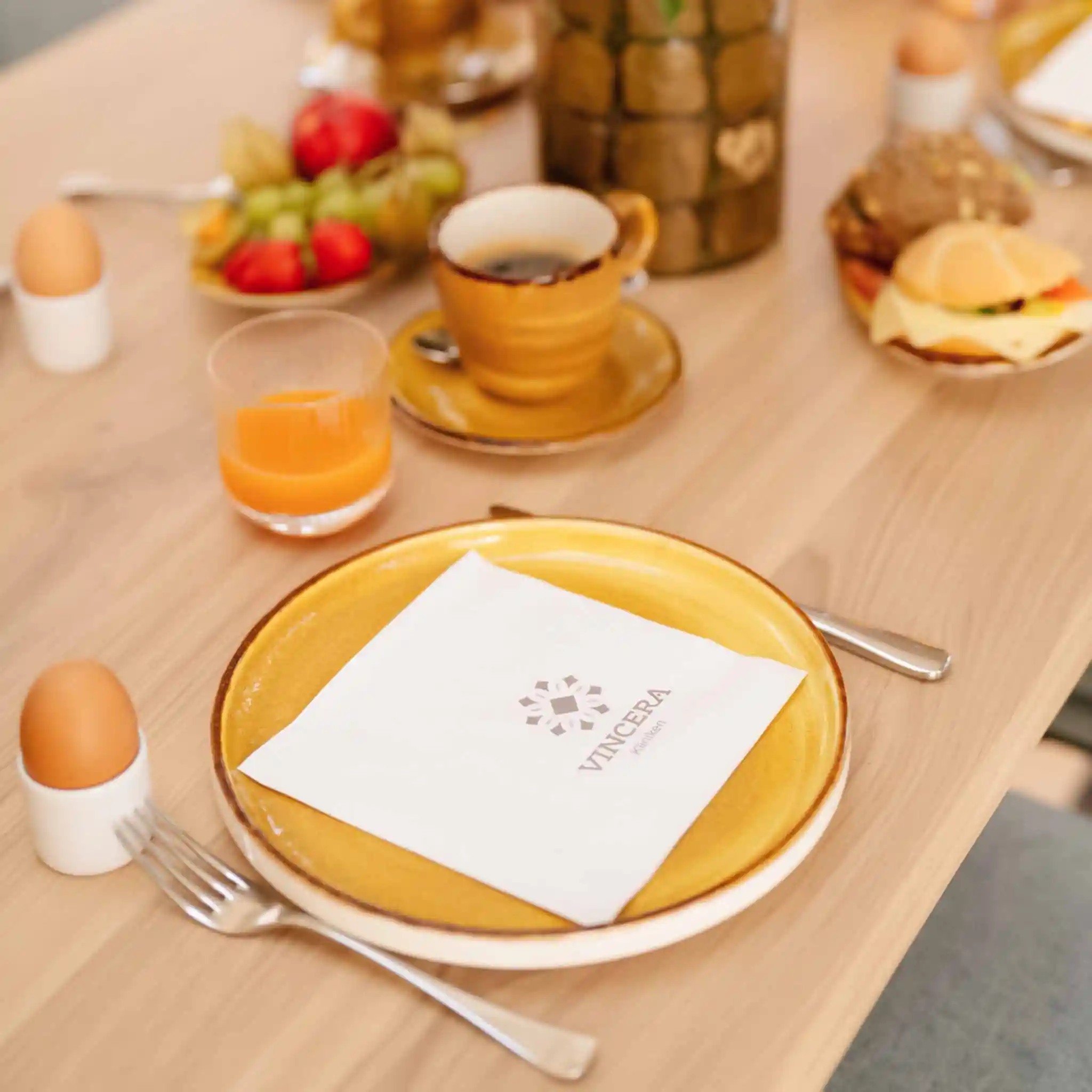 Eine mit dem Vincera Logo bedruckte Serviette liegt auf einem gelben Frühstücksteller.