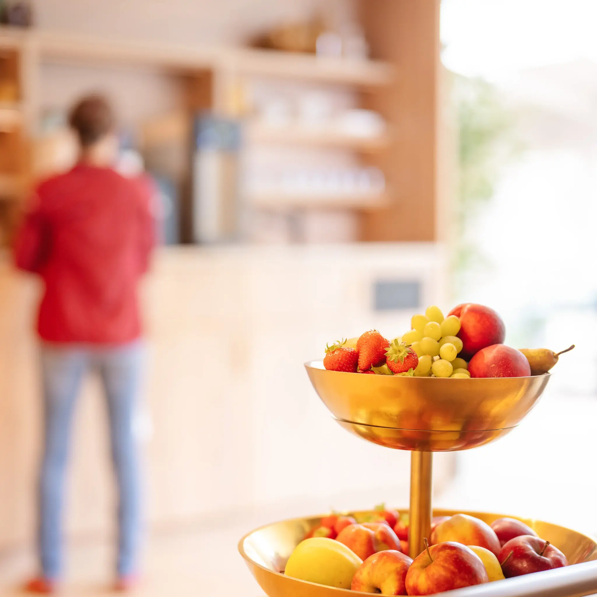 Eine Etagere voll mit frischem Obst. Im Hintergrund sieht man verschwommen eine Person, die sich einen Kaffee an einer Kaffeemaschine holt.