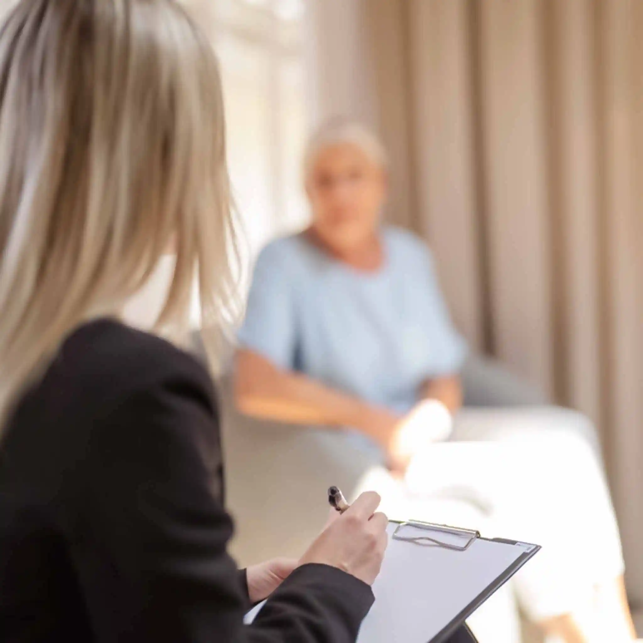 Eine Therapeutin notiert etwas auf ihrem Klemmbrett, während sie mit einer Patientin spricht.