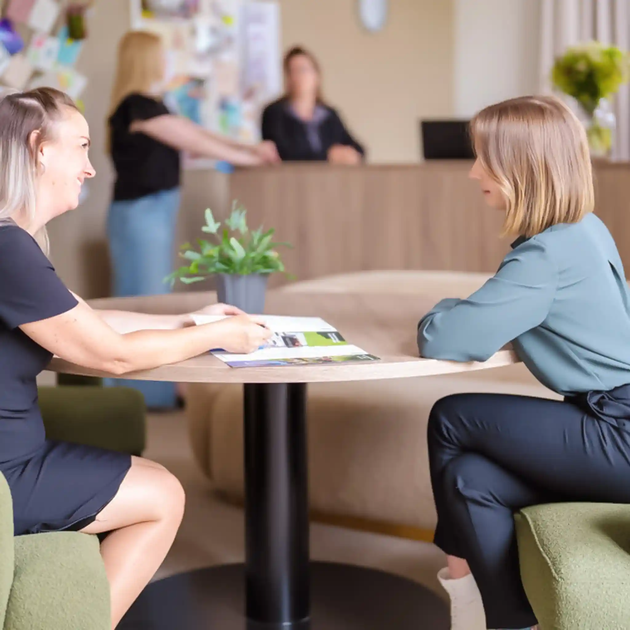 Eine Patientenmanagerin zeigt einer Patientin einige Infomaterialien. Sie sitzen sich an einem Tisch gegenüber.