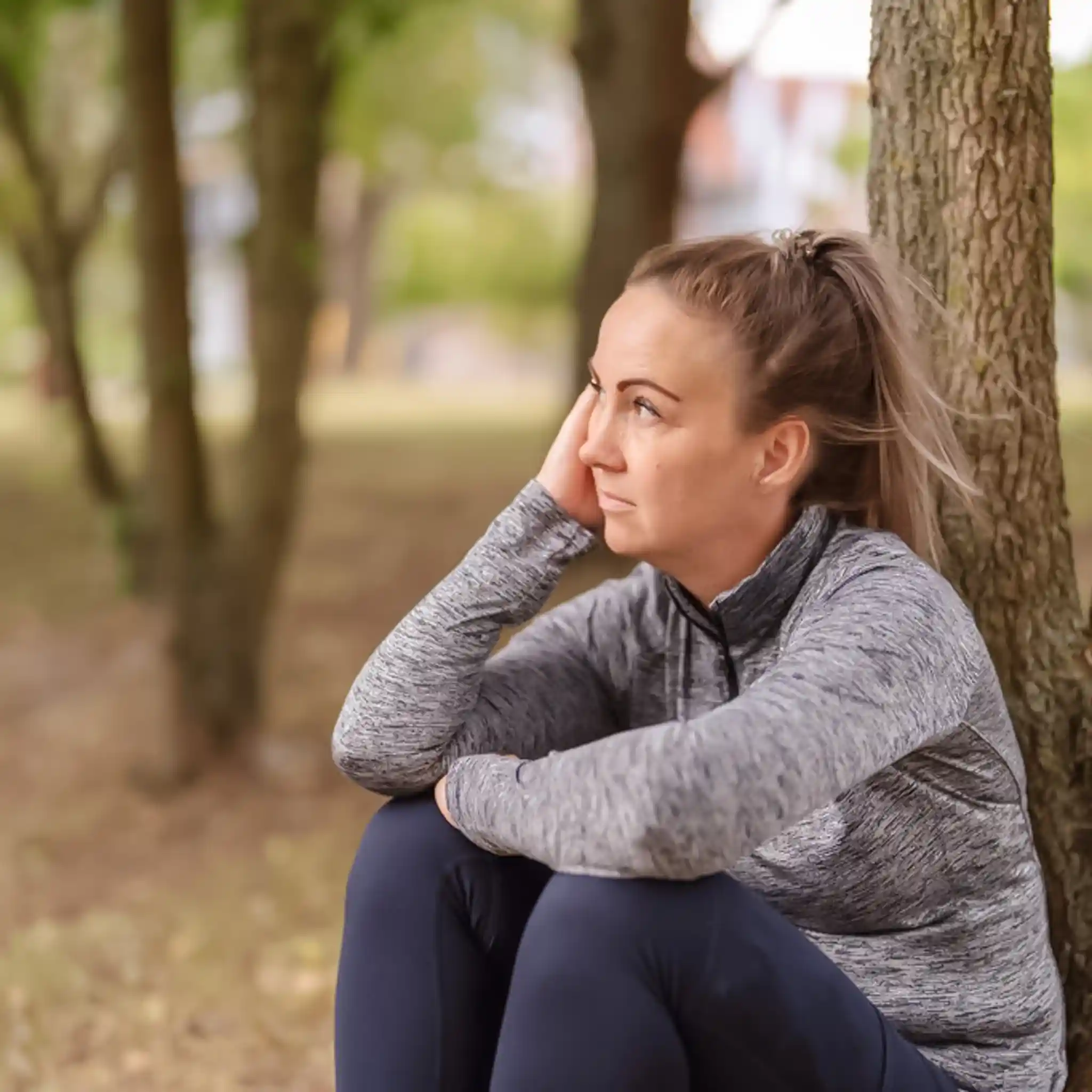 Eine Patientin einer Psychiatrie sitzt an einem Baum gelehnt auf dem Boden.