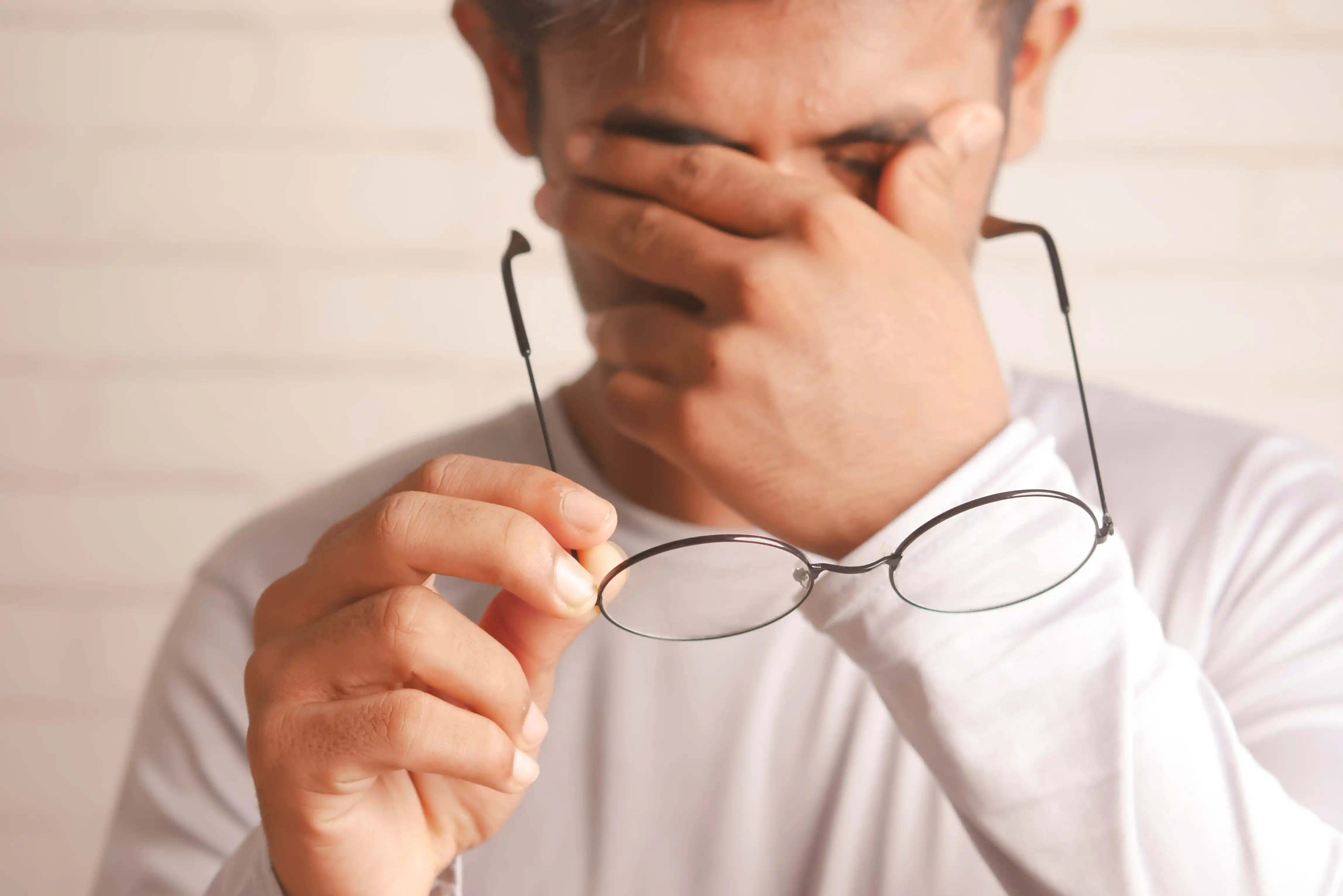 Mann in weißem Hemd hält Brille und reibt sich die Augen mit der anderen Hand.