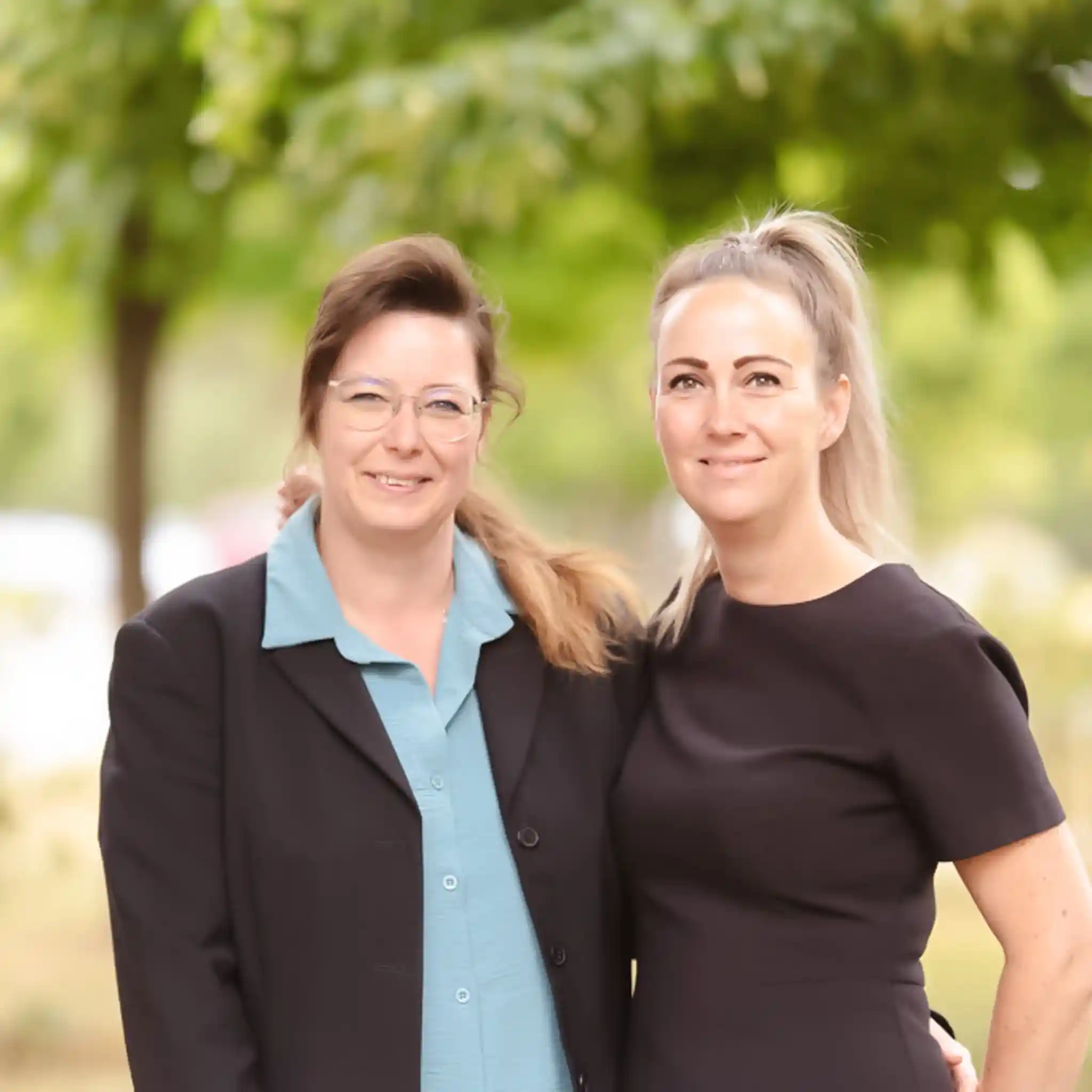 Zwei lächelnde Frauen, die nebeneinander stehen, eine mit Brille und schwarzem Blazer über einem blauen Hemd, die andere in einem schwarzen Kleid, im Freien vor grünem, unscharfem Hintergrund.