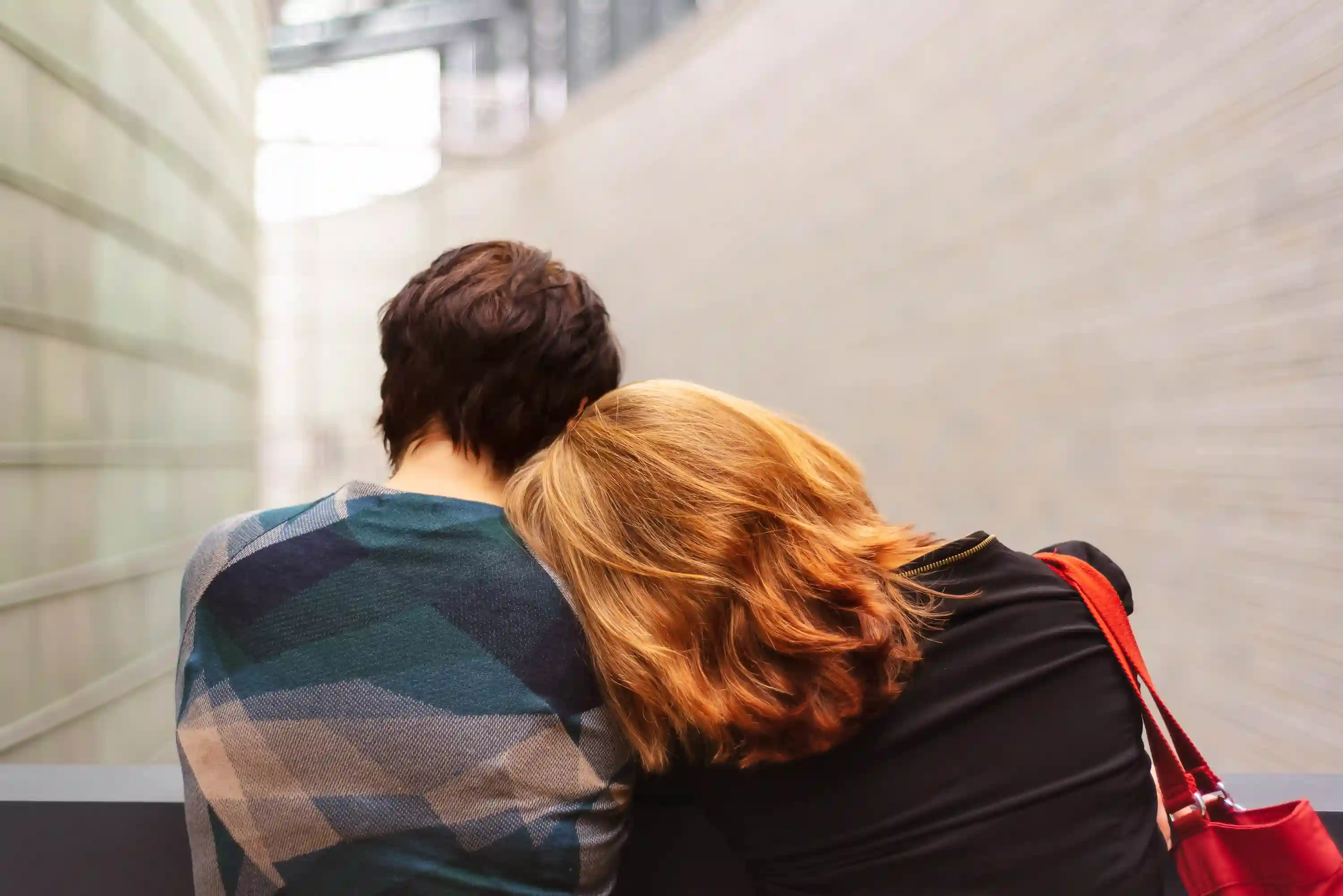 Zwei Personen von hinten, eine mit kurzen braunen Haaren, die andere mit schulterlangen blonden Haaren, die sich an die Schulter der anderen lehnt, in einem hellen, schmalen Gang.